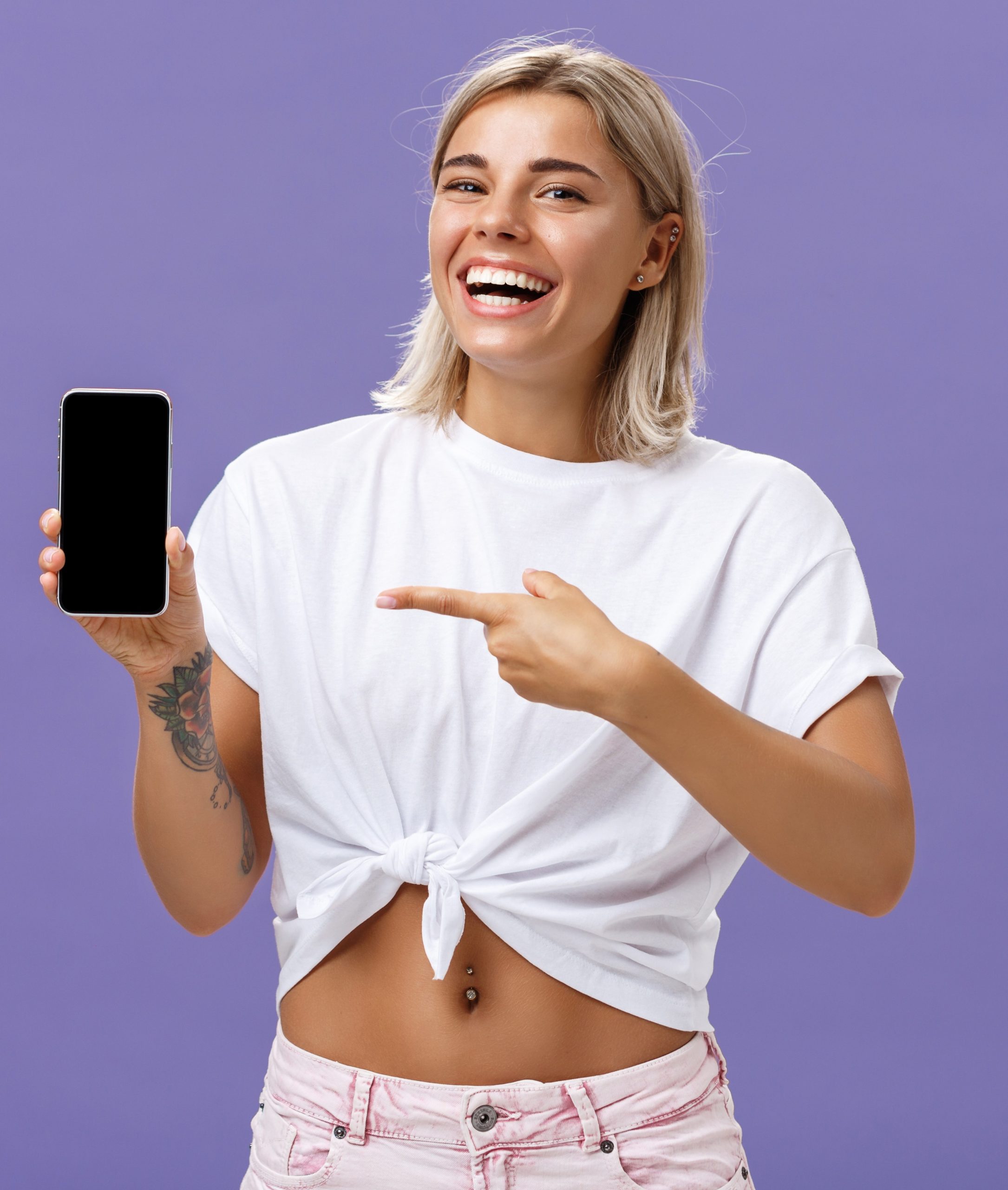 satisfied-happy-beautiful-tanned-woman-with-blond-hairstyle-tattoos-holding-smartphone-showing-device-screen-pointing-gadget-with-index-finger-smiling-camera-purple-wall